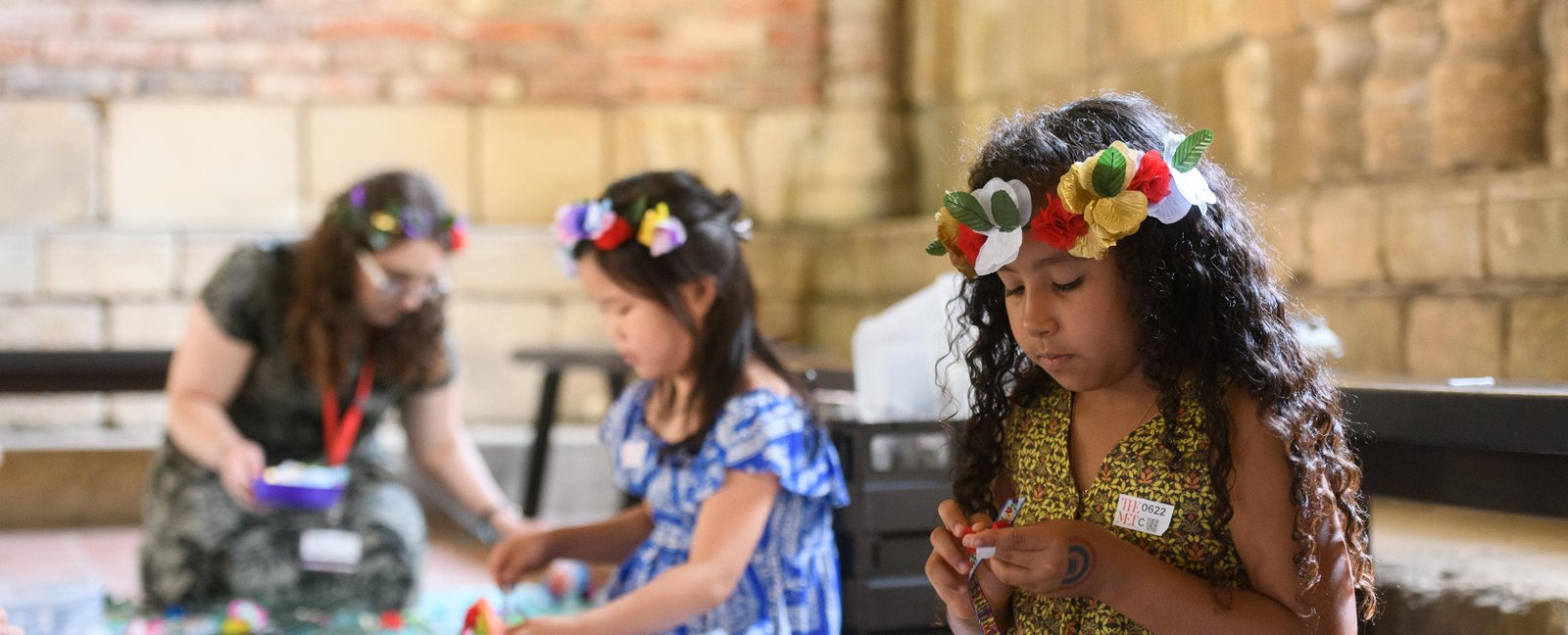 Two girls, wearing paper flower garlands in their hair engage in arts and crafts within a stone walled gallery.