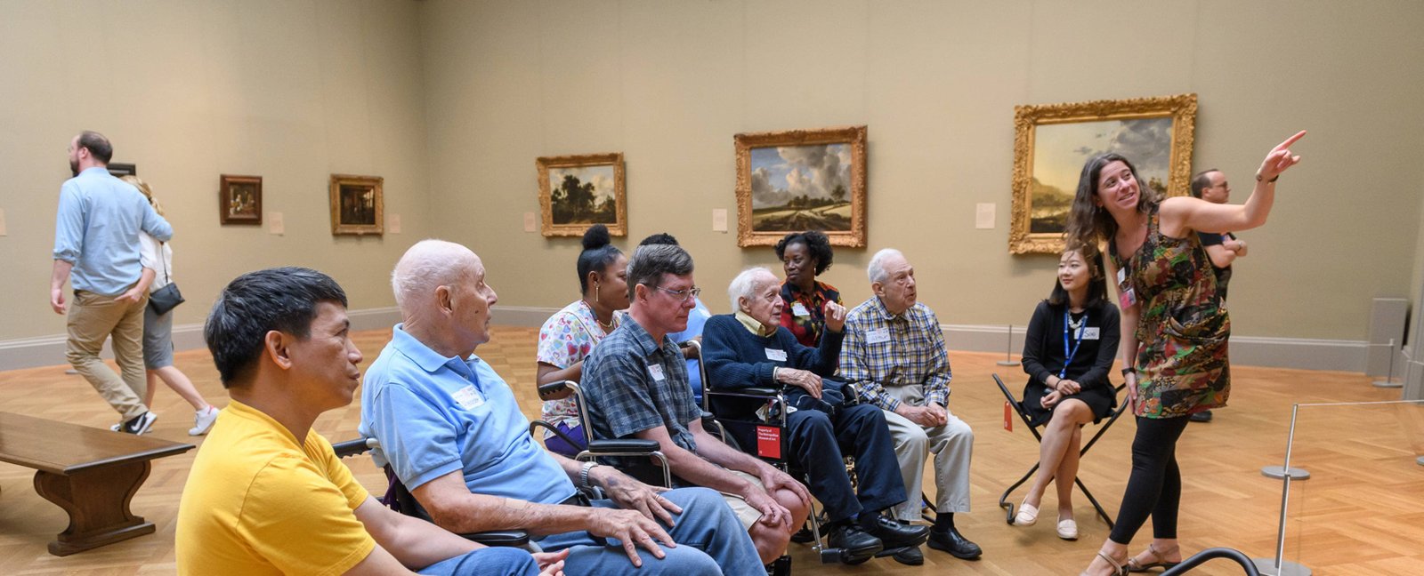 A group of visitors sit inside a gallery facing a Met educator who is pointing to a painting out of our view.