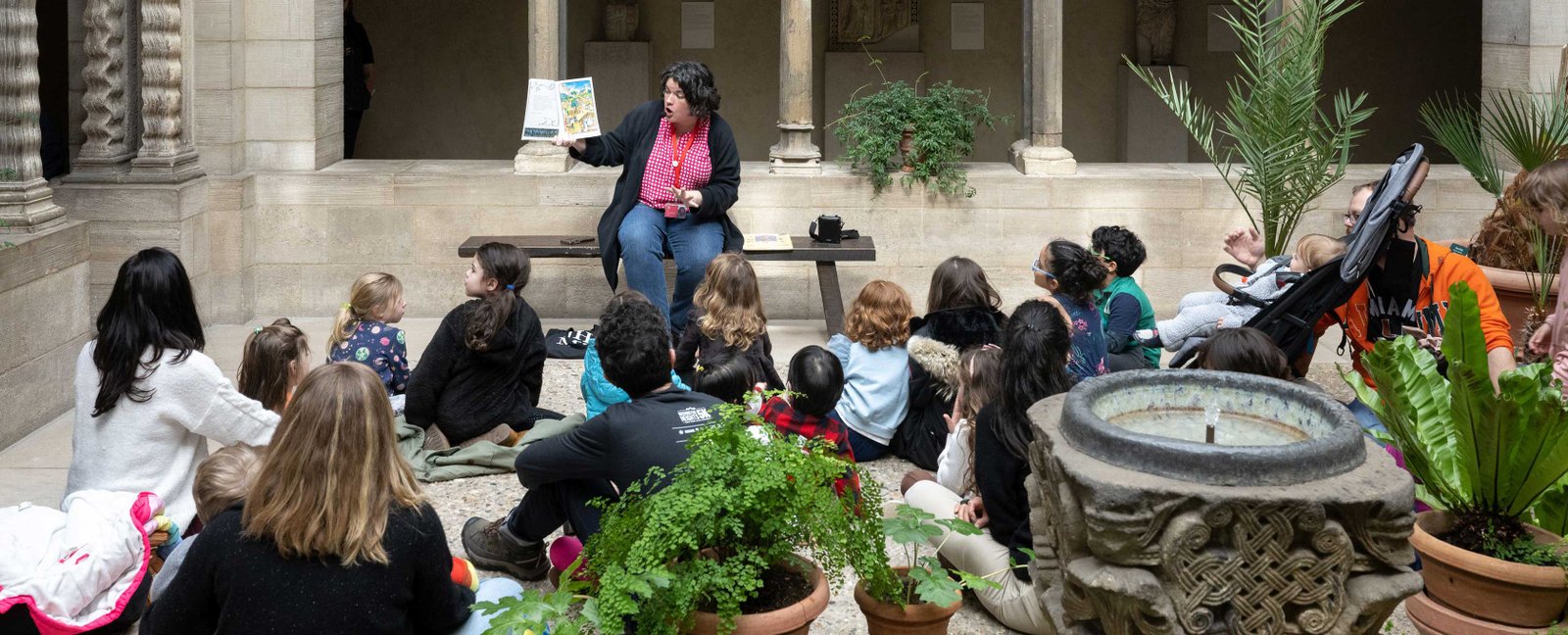 A Met educator reads a picture book to a group of children seated on the floor. They sit in a room filled with potted plants and surrounded by arched columns.